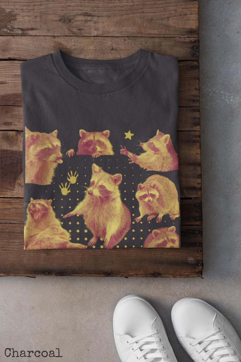 K&ouml;nnte beinhalten: Anthrazitfarbenes T-Shirt mit einem Design aus gelben und rosa Waschb&auml;ren in verschiedenen Posen. Das Design umfasst einen Stern und Handabdr&uuml;cke auf dunklem Hintergrund. Das Wort &bdquo;Charcoal&ldquo; ist unten aufgedruckt.