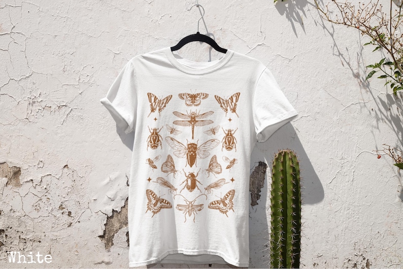May include: White t-shirt with a brown insect design. The illustration features butterflies, dragonflies, and beetles. The t-shirt is suspended on a black hanger against a textured white wall.