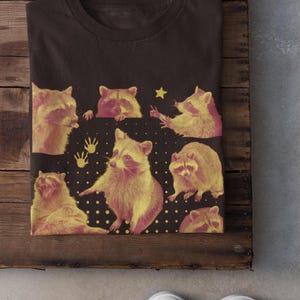 K&ouml;nnte beinhalten: Dunkelbraunes T-Shirt mit einem Design aus gelben und rosa Waschb&auml;ren. Das Design enth&auml;lt einen Stern und Handabdr&uuml;cke auf einem schwarzen Hintergrund mit wei&szlig;en Punkten. Der Text "Dark Chocolate" ist sichtbar.