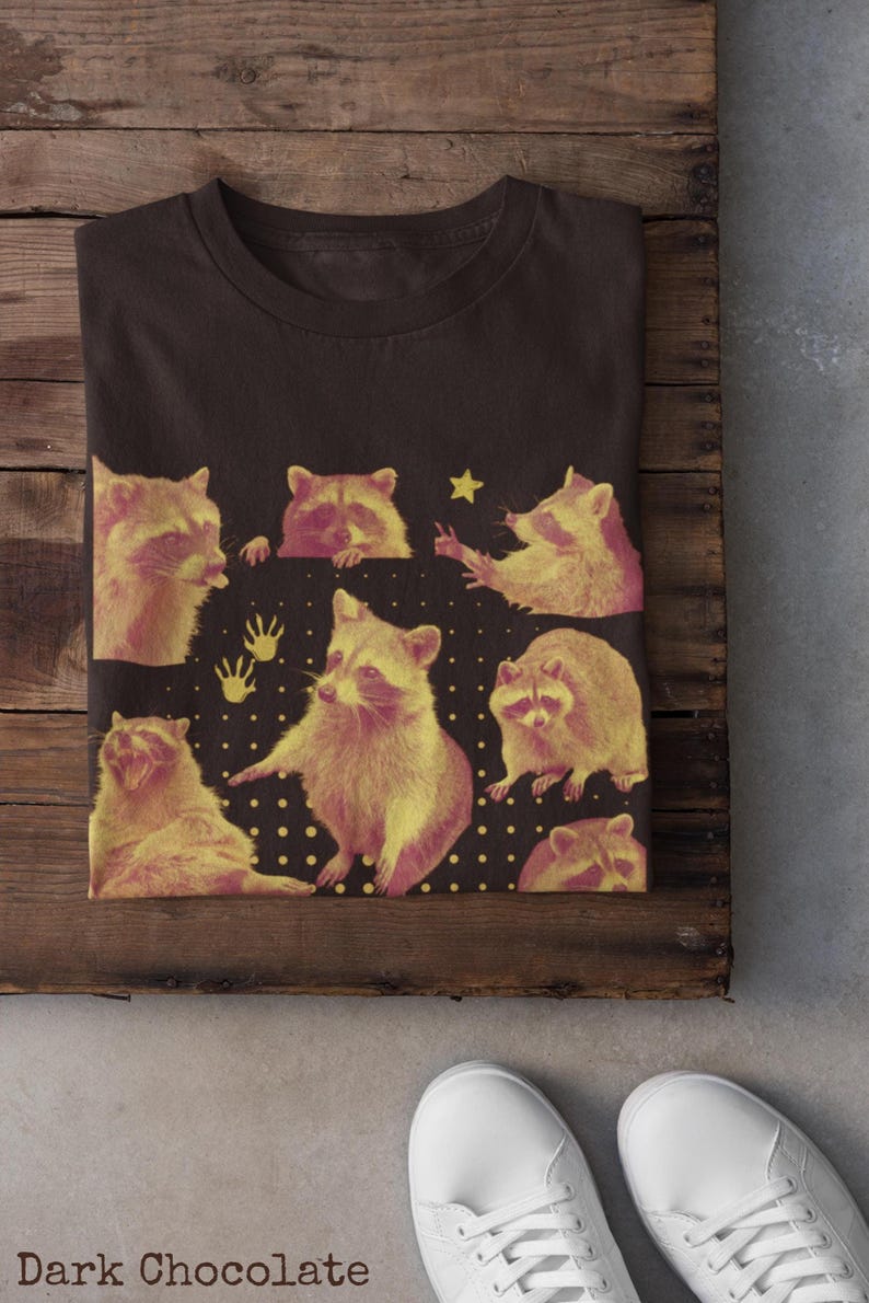 K&ouml;nnte beinhalten: Dunkelbraunes T-Shirt mit einem Design aus gelben und rosa Waschb&auml;ren. Das Design enth&auml;lt einen Stern und Handabdr&uuml;cke auf einem schwarzen Hintergrund mit wei&szlig;en Punkten. Der Text "Dark Chocolate" ist sichtbar.