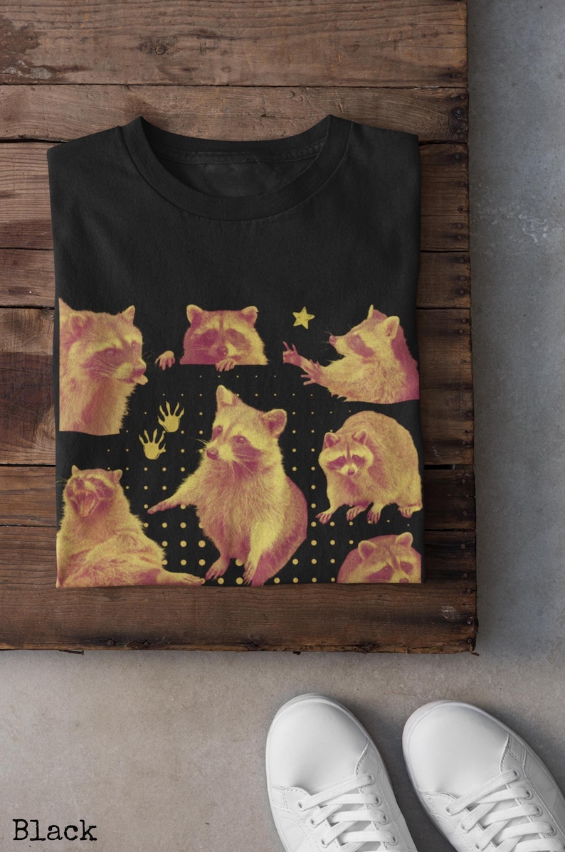 K&ouml;nnte beinhalten: Schwarzes T-Shirt mit einem sich wiederholenden Muster von Cartoon-Waschb&auml;ren in Gelb- und Rosat&ouml;nen. Das Design enth&auml;lt einen Stern und Handabdr&uuml;cke auf schwarzem Hintergrund. Das Wort "Black" steht unten.