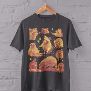 K&ouml;nnte beinhalten: Anthrazitfarbenes T-Shirt mit einer Collage von Waschb&auml;ren in verschiedenen Posen mit gelben und roten Akzenten. Das Design beinhaltet Sterne und Handabdr&uuml;cke. Das Wort "Charcoal" ist unten aufgedruckt.