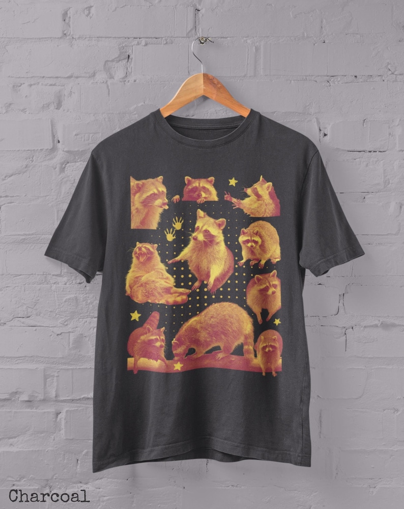 K&ouml;nnte beinhalten: Anthrazitfarbenes T-Shirt mit einer Collage von Waschb&auml;ren in verschiedenen Posen mit gelben und roten Akzenten. Das Design beinhaltet Sterne und Handabdr&uuml;cke. Das Wort "Charcoal" ist unten aufgedruckt.