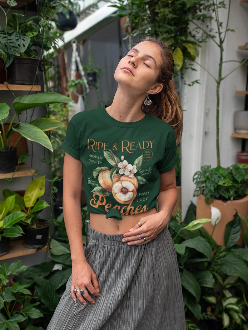 May include: Dark green t-shirt with the text "Ripe & Ready" and a peach graphic. The shirt features a crew neck and short sleeves. The design includes peaches, flowers, and the words "Hidden Valley" and "Sweet Juicy Peaches".