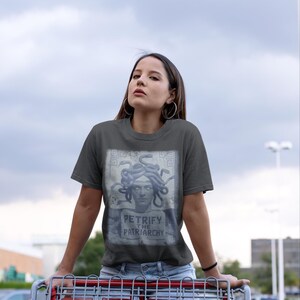 May include: Dark gray t-shirt featuring a Medusa graphic with the text "Petrify the Patriarchy." The shirt is made of a soft material. The person is wearing the shirt while holding a shopping cart.