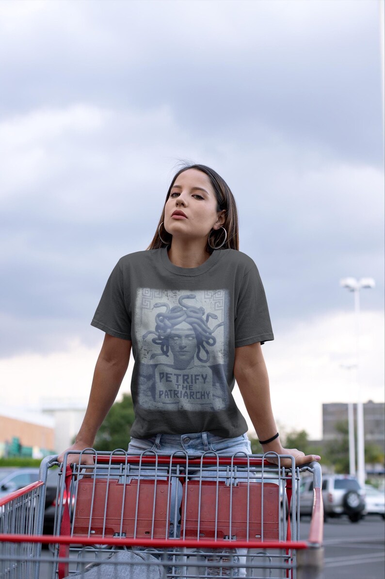 May include: Dark gray t-shirt featuring a Medusa graphic with the text "Petrify the Patriarchy." The shirt is made of a soft material. The person is wearing the shirt while holding a shopping cart.
