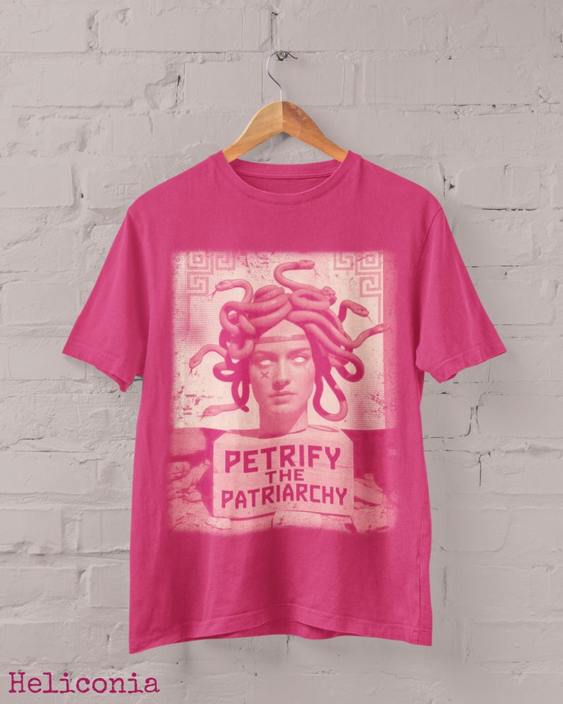 May include: A pink t-shirt with a graphic of Medusa, depicted with snakes for hair, and the text "PETRIFY THE PATRIARCHY". The design is in a rectangular frame. The shirt is hanging on a wooden hanger.