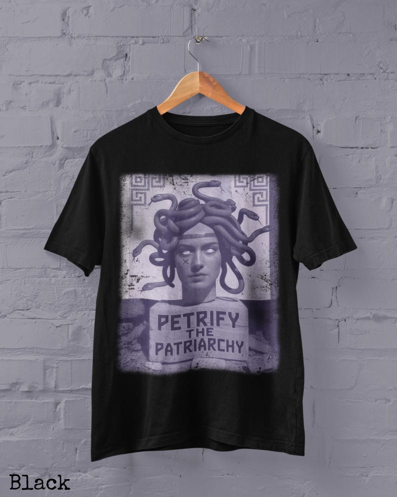 May include: Black t-shirt with a graphic of Medusa, depicted with snakes for hair, a white face, and the text "PETRIFY THE PATRIARCHY". The design is in purple and white. The t-shirt is displayed on a wooden hanger.