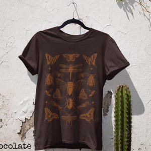Pu&ograve; includere: T-shirt color cioccolato fondente con un design arancione di insetti e farfalle. Il design include vari insetti e farfalle. Il testo "Dark Chocolate" &egrave; in basso nell'immagine.