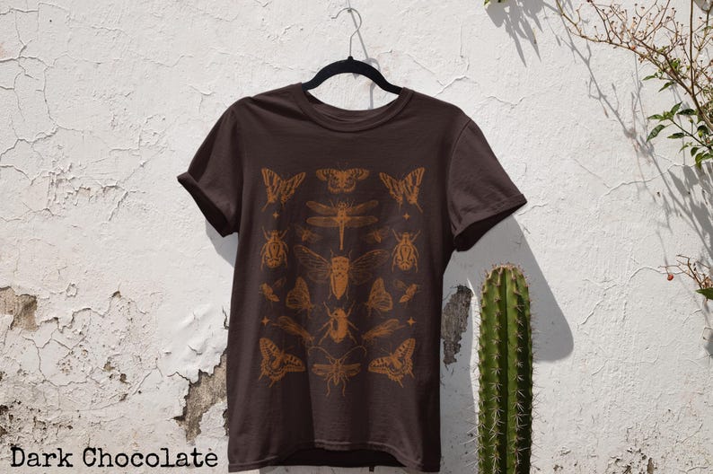 Pu&ograve; includere: T-shirt color cioccolato fondente con un design arancione di insetti e farfalle. Il design include vari insetti e farfalle. Il testo "Dark Chocolate" &egrave; in basso nell'immagine.