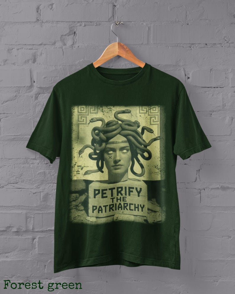 May include: Forest green t-shirt with a graphic of Medusa, her hair made of snakes, and the text "Petrify the Patriarchy." The design is in a rectangular frame. The shirt hangs on a wooden hanger against a grey brick wall.