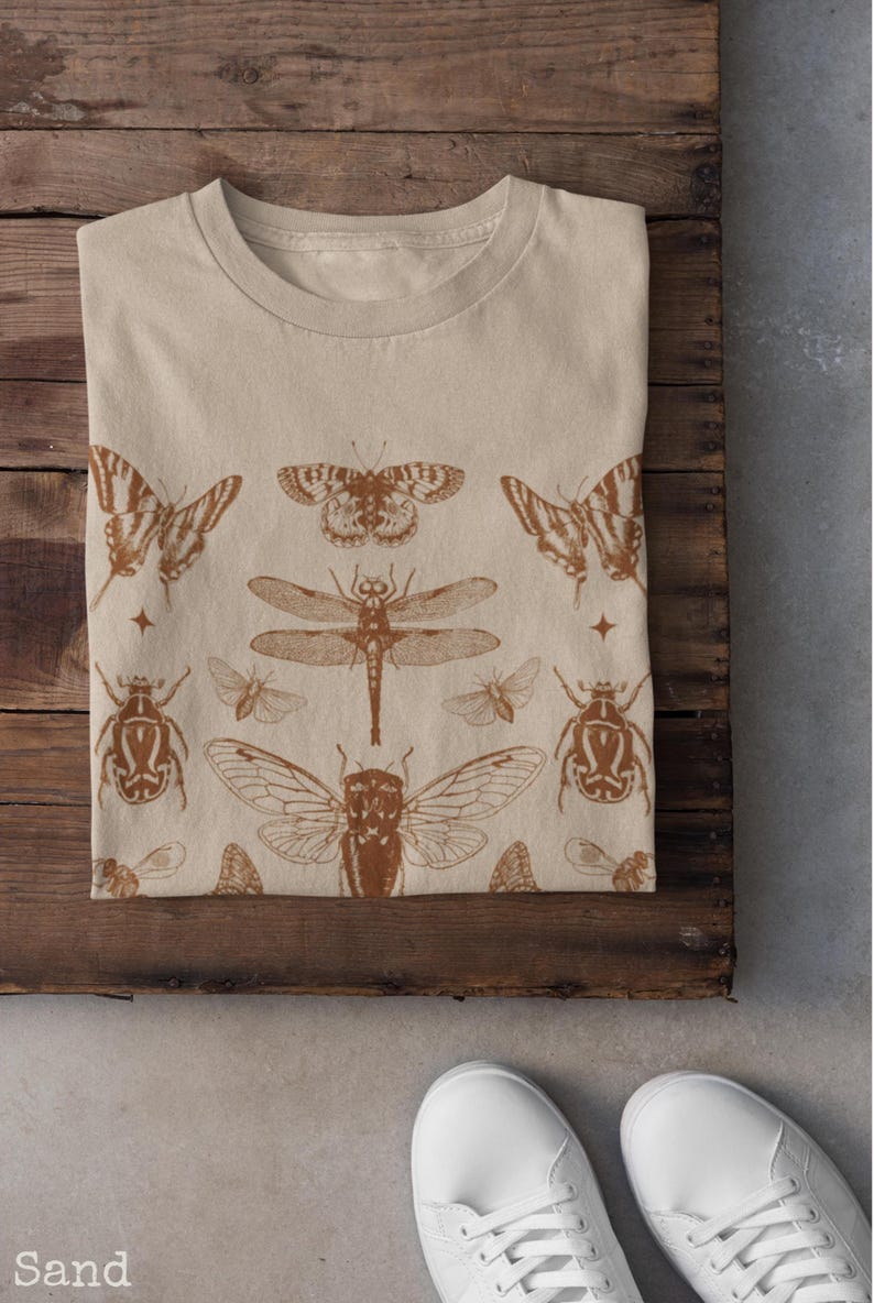 Pu&ograve; includere: T-shirt beige con un disegno di insetti marroni. Il design include farfalle, una libellula, scarabei e altri insetti. La maglietta &egrave; piegata su una superficie di legno, con scarpe da ginnastica bianche.