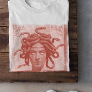 May include: White t-shirt featuring a Medusa graphic in shades of red. The design depicts Medusa's face with snakes for hair, set against a textured background. The shirt is displayed on a wooden surface.