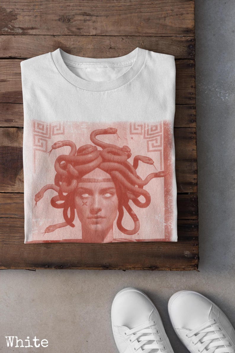May include: White t-shirt featuring a Medusa graphic in shades of red. The design depicts Medusa's face with snakes for hair, set against a textured background. The shirt is displayed on a wooden surface.