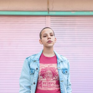 May include: A person wearing a pink t-shirt with a graphic of Medusa and the text "PETRIFY THE PATRIARCHY". The person is also wearing a light blue denim jacket. The background is a pink wall.