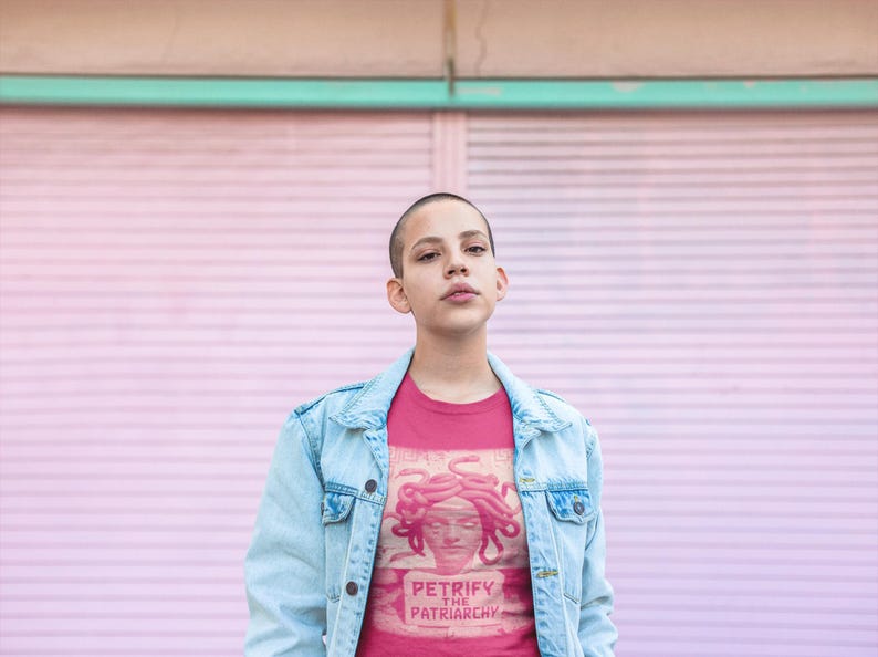 May include: A person wearing a pink t-shirt with a graphic of Medusa and the text "PETRIFY THE PATRIARCHY". The person is also wearing a light blue denim jacket. The background is a pink wall.