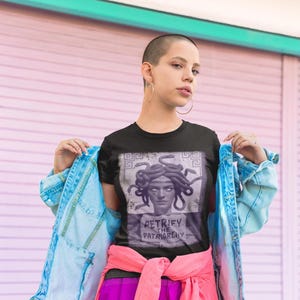 May include: Black t-shirt featuring a graphic of Medusa with the text "PETRIFY THE PATRIARCHY". The shirt is paired with a light blue denim jacket, pink shirt tied around the waist, and purple pants. The background is a pink and white wall.