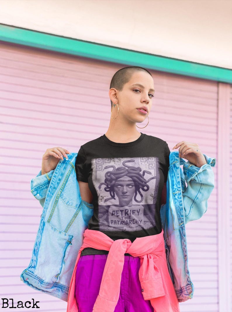 May include: Black t-shirt featuring a graphic of Medusa with the text "PETRIFY THE PATRIARCHY". The shirt is paired with a light blue denim jacket, pink shirt tied around the waist, and purple pants. The background is a pink and white wall.