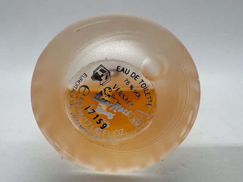 Op de afbeelding: Close-up van de bodem van een parfumfles, met een etiket met de tekst "EAU DE TOILETTE" en "VERSACE". De fles bevat een lichtoranje vloeistof. Het etiket geeft ook een volume van ongeveer 50 ml aan.
