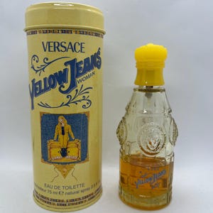 May include: A yellow cylindrical tin and a perfume bottle. The tin reads "VERSACE Yellow Jeans Woman" with a blue denim-like background. The perfume bottle is clear with a yellow cap and contains a golden liquid. The bottle has the words "Yellow Jeans" in blue.