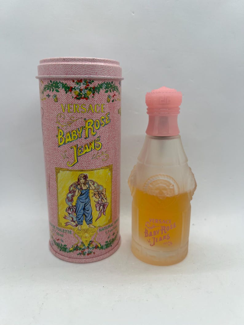 Op de afbeelding: Een roze cilindrische blik en een parfumfles. De blik bevat de tekst "Versace Baby Rose Jeans" en een afbeelding van een vrouw. De parfumfles is helder met een roze dop en bevat een gele vloeistof.