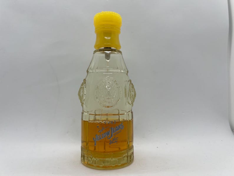 May include: A clear glass perfume bottle with a yellow cap. The bottle has a rectangular shape with embossed details and contains a golden-coloured liquid. The words "Yellow Jeans" are printed in blue on the front.