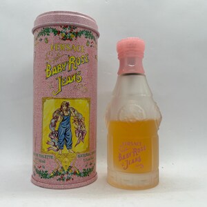 Op de afbeelding: Een vintage Versace Baby Rose Jeans parfumflesje en bijpassende cilindrische container. De fles heeft een roze dop en bevat een gele vloeistof. De container is roze met bloemaccenten en een label met een vrouw in een blauwe overall.