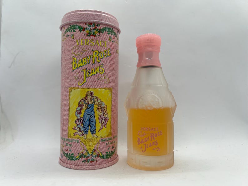 Op de afbeelding: Een vintage Versace Baby Rose Jeans parfumflesje en bijpassende cilindrische container. De fles heeft een roze dop en bevat een gele vloeistof. De container is roze met bloemaccenten en een label met een vrouw in een blauwe overall.