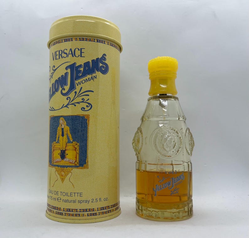 May include: Versace Yellow Jeans Woman eau de toilette, presented in a bottle with a yellow cap and a cylindrical container. The container displays the Versace logo and a denim-themed illustration. The bottle contains 75 ml of perfume.