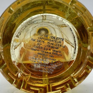 May include: Close-up of the base of a perfume bottle filled with a golden liquid. The clear glass base features etched details and text including "AU DE TOILETTE VAPORISATEUR" and "75 ml - 2.5 fl. oz."