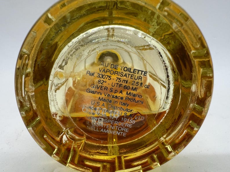 May include: Close-up of the base of a perfume bottle filled with a golden liquid. The clear glass base features etched details and text including "AU DE TOILETTE VAPORISATEUR" and "75 ml - 2.5 fl. oz."