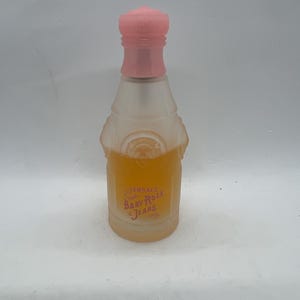 Op de afbeelding: Een kleine, matglazen parfumfles met een roze dop. De fles bevat een licht amberkleurige vloeistof en heeft de tekst "Versace Baby Rose Jeans" in roze letters. De fles heeft een unieke vorm en design.