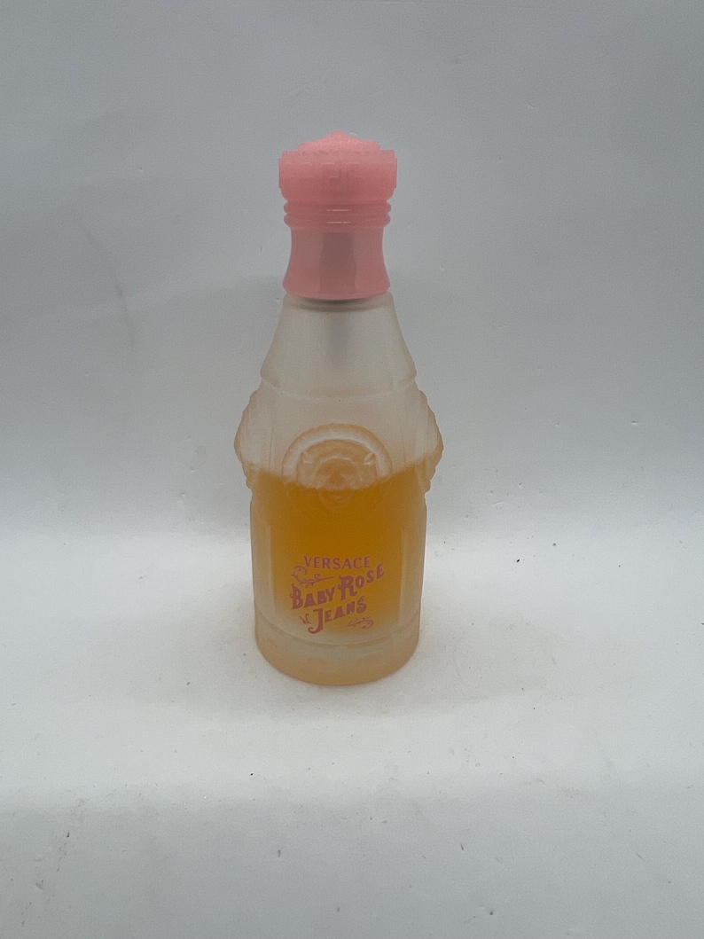 Op de afbeelding: Een kleine, matglazen parfumfles met een roze dop. De fles bevat een licht amberkleurige vloeistof en heeft de tekst "Versace Baby Rose Jeans" in roze letters. De fles heeft een unieke vorm en design.