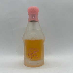 Op de afbeelding: Een heldere glazen parfumfles met een roze dop en een lichtoranje vloeistof erin. De fles heeft een label met de tekst "Versace Baby Rose Jeans". De fles heeft de vorm van een champagnefles.
