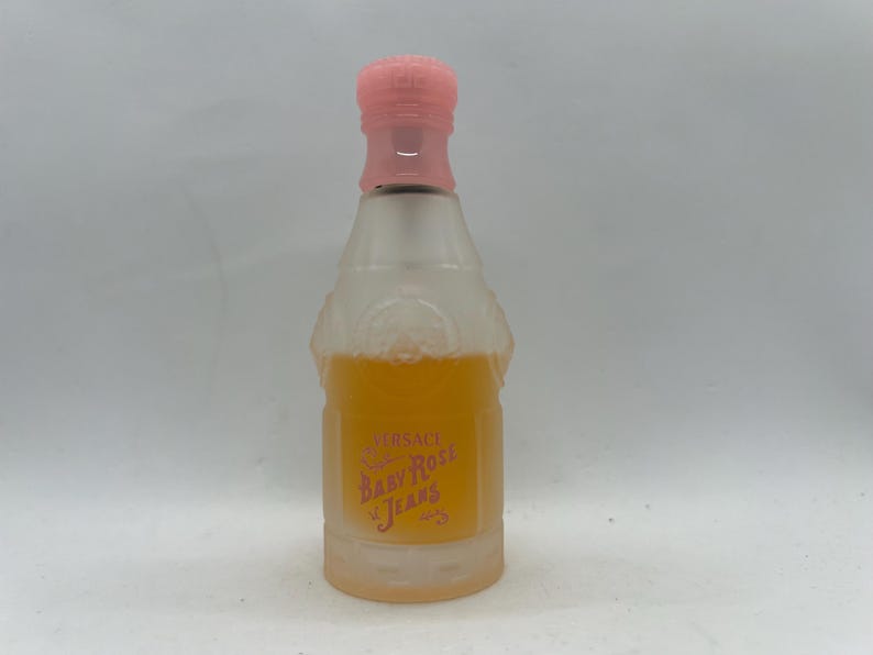 Op de afbeelding: Een heldere glazen parfumfles met een roze dop en een lichtoranje vloeistof erin. De fles heeft een label met de tekst "Versace Baby Rose Jeans". De fles heeft de vorm van een champagnefles.