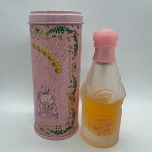Op de afbeelding: Een vintage parfumfles en bijpassende cilindrische container. De fles heeft een roze dop en een heldere, matte body met gele vloeistof erin. De container is roze met een denimachtig patroon en een rugzakillustratie.