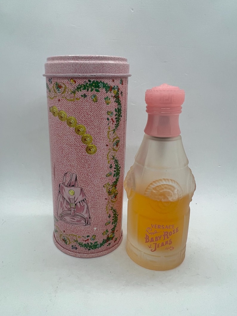 Op de afbeelding: Een vintage parfumfles en bijpassende cilindrische container. De fles heeft een roze dop en een heldere, matte body met gele vloeistof erin. De container is roze met een denimachtig patroon en een rugzakillustratie.