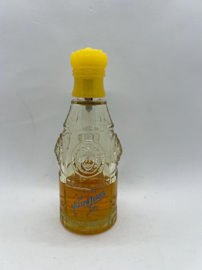 May include: A clear glass perfume bottle with a yellow cap. The bottle has a textured design and contains a golden-coloured liquid. The words "Yellow Jeans" are printed on the front of the bottle.