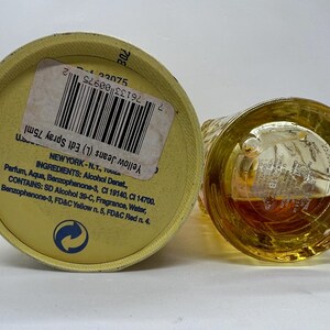 May include: A yellow perfume bottle and its cap are shown. The cap, with a label, is on its side, and the bottle is upright. The label reads "Yellow Jeans Edt Spray 75ml." The bottle's contents are amber-colored.