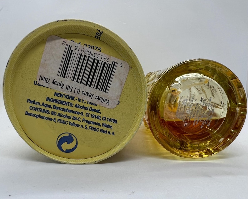 May include: A yellow perfume bottle and its cap are shown. The cap, with a label, is on its side, and the bottle is upright. The label reads "Yellow Jeans Edt Spray 75ml." The bottle's contents are amber-colored.