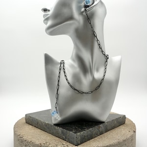 May include: A pair of clear and light blue earplugs connected by a long, thin black metal chain. The chain features elongated oval links and is draped over a silver-colored mannequin head and neck display piece to show the length and style.