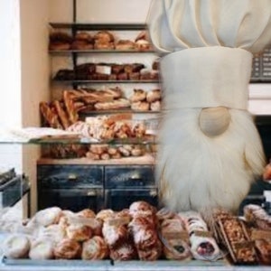 May include: A white chef hat gnome figurine standing in front of a bakery display case filled with various pastries and breads.