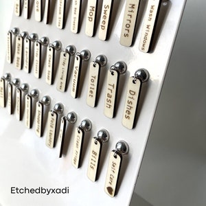 May include: A white board with wooden tags attached to it with silver tacks. The tags are labeled with various household chores, such as "Make Bed", "Dust", "Stove", "Wash Windows", "Dishes", "Sweep", "Mop", "Toilet", "Trash", "Clean Fridge", "Vacuum", "Wash Bed Sheets", "Tidy Living Area", "Fold Laundry", "Bathroom Sink", "Check Soap", "Tidy Closet", "Laundry", "Make Beds", "Shopping", "Self Care", and "Bills". The board is labeled "Etchedbyxadi" at the bottom.