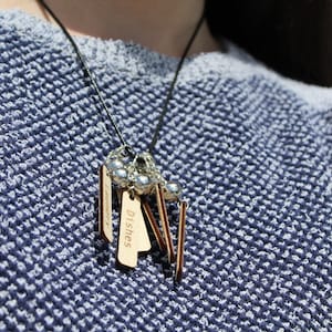 May include: A black leather necklace with wooden tags that say "Dishes" and "Laundry". The necklace has silver beads and is worn by a person wearing a blue and white sweater.