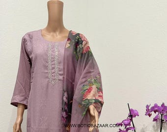 May include: A lilac-colored salwar kameez set with a matching dupatta. The tunic and pants have a subtle vertical stripe pattern. The dupatta features a floral print with pink and purple flowers. The neckline is embellished with silver beads. The website address is visible.