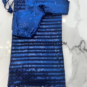 May include: A blue sequined saree with horizontal stripes and a matching blouse. The saree has a decorative border with gold accents. The blouse is also covered in blue sequins. The saree is folded and displayed on a white marble surface.