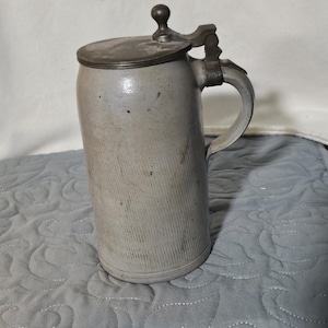 May include: A gray stoneware beer stein with a hinged metal lid and handle. The cylindrical body has vertical grooves. The lid is topped with a small, round knob. The handle is attached to the side of the stein.