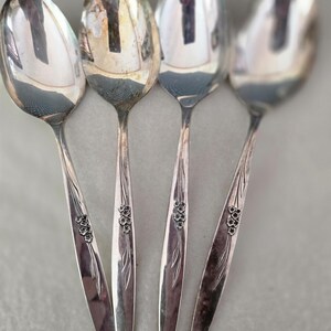 May include: Four silver-toned metal spoons arranged side by side. Each spoon features a polished oval bowl and a tapered handle decorated with a small, embossed floral motif consisting of five petals and slender, etched leaf patterns.