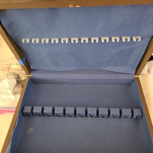 May include: A rectangular brown wooden storage box with a hinged lid, featuring a light blue fabric-lined interior. The lid contains a row of white plastic hooks, while the base includes a row of light blue fabric-covered slots for organizing.