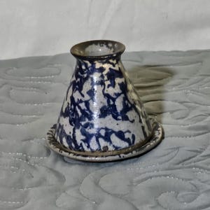 May include: A ceramic vase with a flared base and a narrow opening. The vase is white with a blue splatter pattern and sits on a matching saucer. The rim of the vase is dark brown.